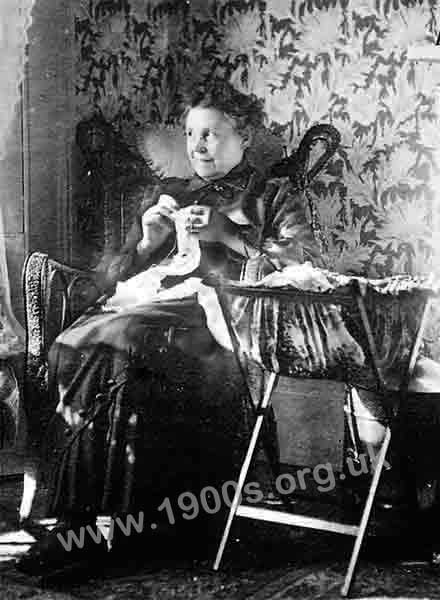 A working class woman sewing, mending and dressmaking in the early 1900s, showing her clothes, her needlework bag and her wickerwork chair.