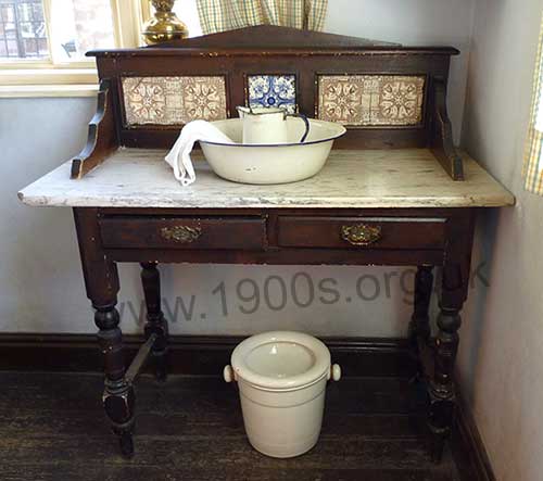 Old washstand, as used in Victorian and Edwardian Britain, with the customary marble top and the matching jug and bowl set. In this photo the jug and bowl set are made of enamel which would have been cheaper than decorated china.